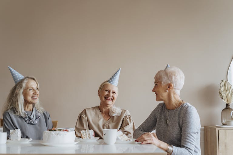 Three senior women enjoy a cozy indoor birthday celebration with party hats and smiles.