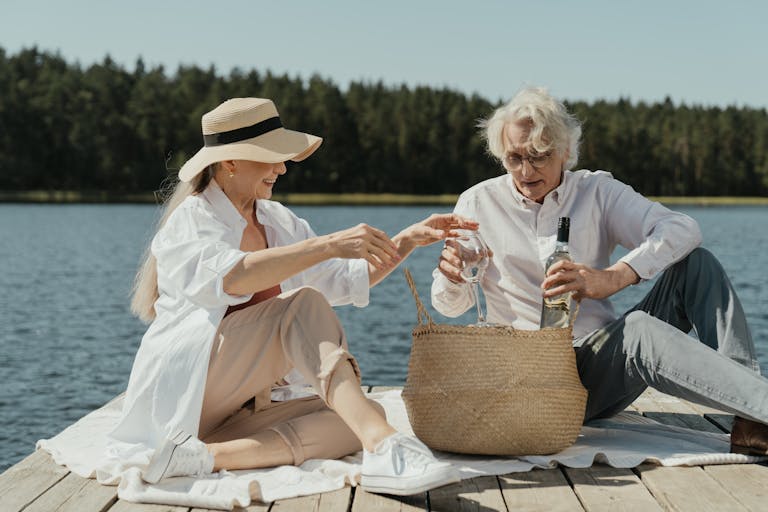 Elderly couple sits on a pier enjoying a picnic by the lake, embracing lifestyle and leisure.